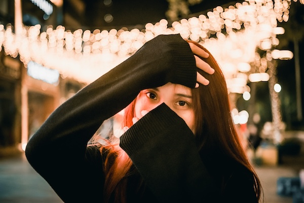 Person covering most of her face with her hands, standing in front of hanging small lights