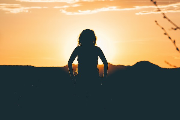 child with their hands on their hips, looking out at a sunset